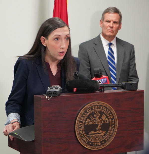 Mary-Margaret Fill, MD, speaks at a lectern before TV cameras as Gov. Bill Lee looks on.